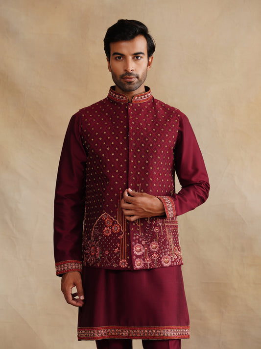 Man wearing a maroon traditional outfit with intricate patterns on a beige background